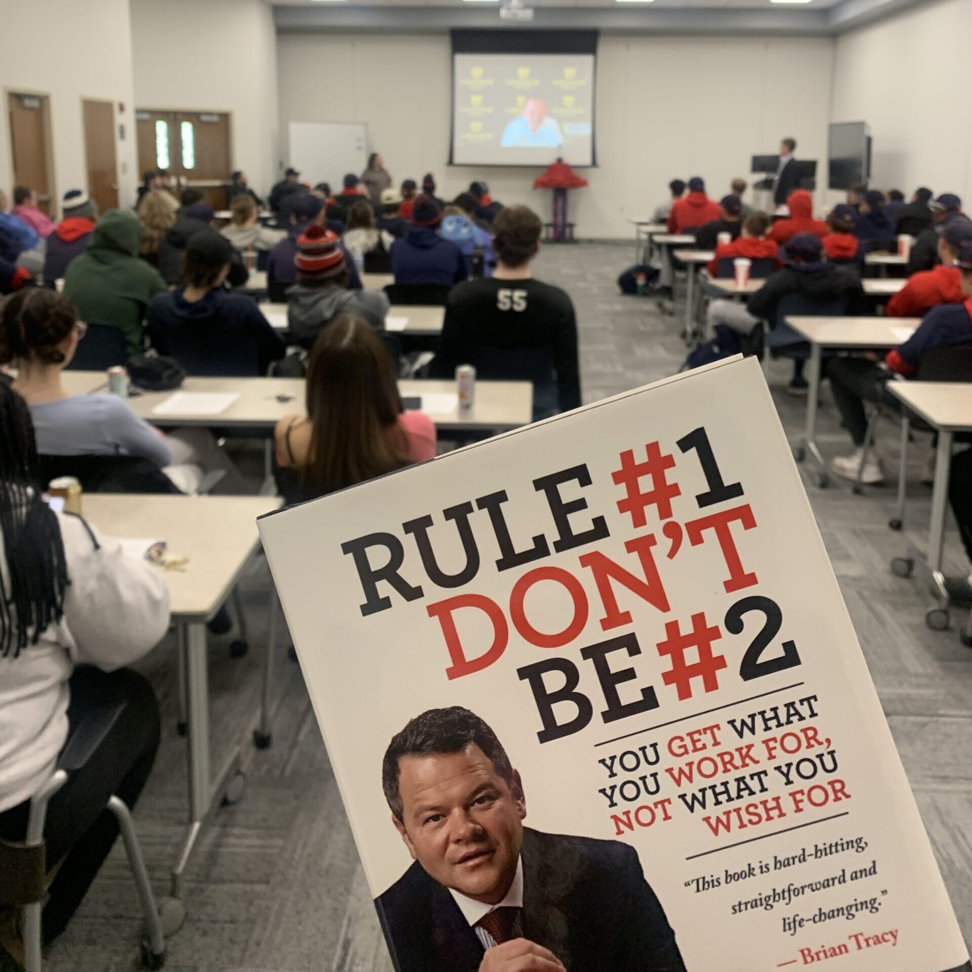 photo of Daniel Milstein's book "Rule number one don't be number two" being held up in front of classroom with Dan on a screen via zoom call.